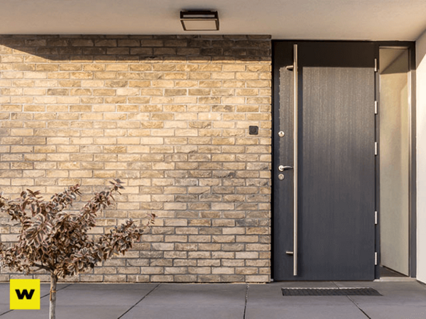 Moderne Haustür aus dunklem Holz in heller Klinkerfassade mit kleinem Baum davor.